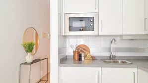 A modern kitchen with white cabinets, a stainless steel sink, and a microwave. There is a small plant on a console table and a wooden cutting board with utensils. - Charming Studio with French Balcony in Upper Málaga, Close to Downtown (Málaga)