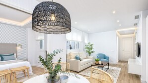 Ett modernt vardagsrum med ett runt matbord, möbler i rotting, en stor svart hänglampa och en soffa med utsikt över utemiljön.