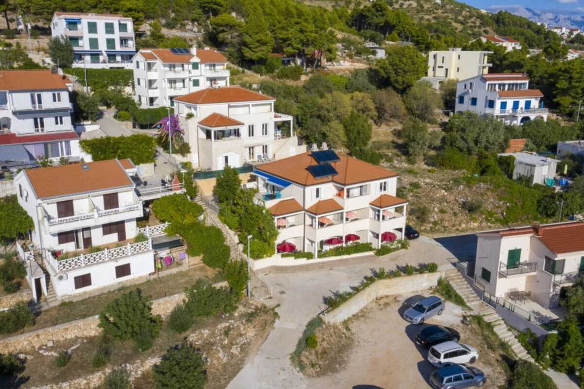 Zona residencial con edificios de varios pisos, algunos con balcones e instalaciones solares, rodeada de vegetación y colinas.
