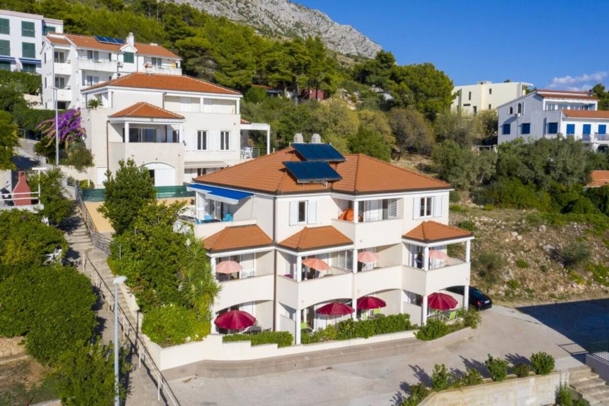 Un edificio de varios pisos con balcones, zona de estacionamiento y un paisaje montañoso al fondo.