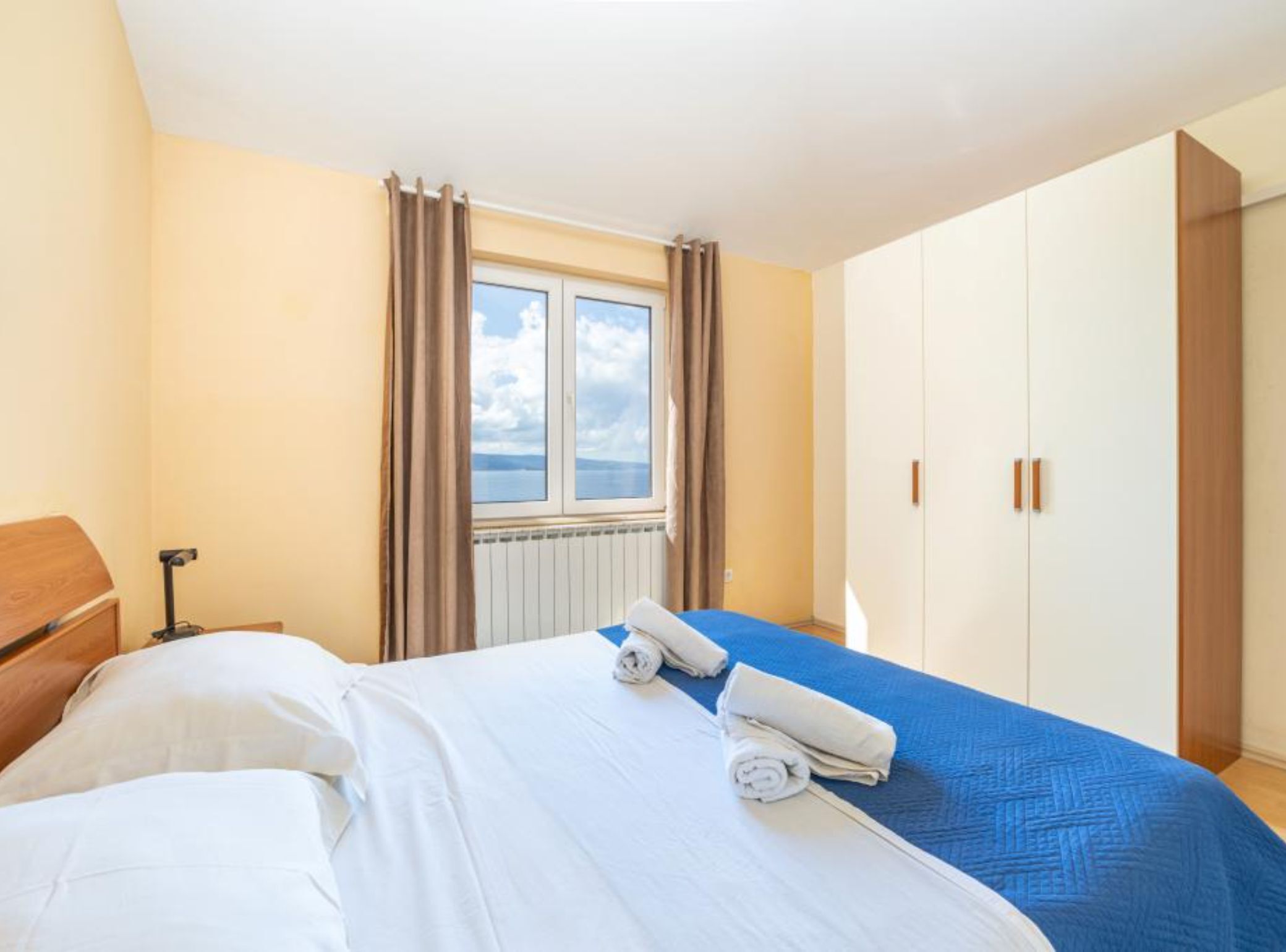 A bedroom with a bed, a window with curtains, a radiator, and a wardrobe.