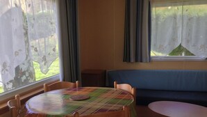 A cozy dining area with a round table covered by a plaid tablecloth, surrounded by wooden chairs. A blue sofa is positioned against the wall, and a window with curtains is visible. - Mobile home with terrace - 4 guests (Saint-Girons)