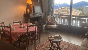 A cozy dining area with a red and white checkered tablecloth, wooden chairs, and a fireplace. A television is mounted on the wall above the fireplace. The room has a large window with a view of mountains and a wooden cabin. - 3-room apartment for 6 people in Combloux (Combloux)