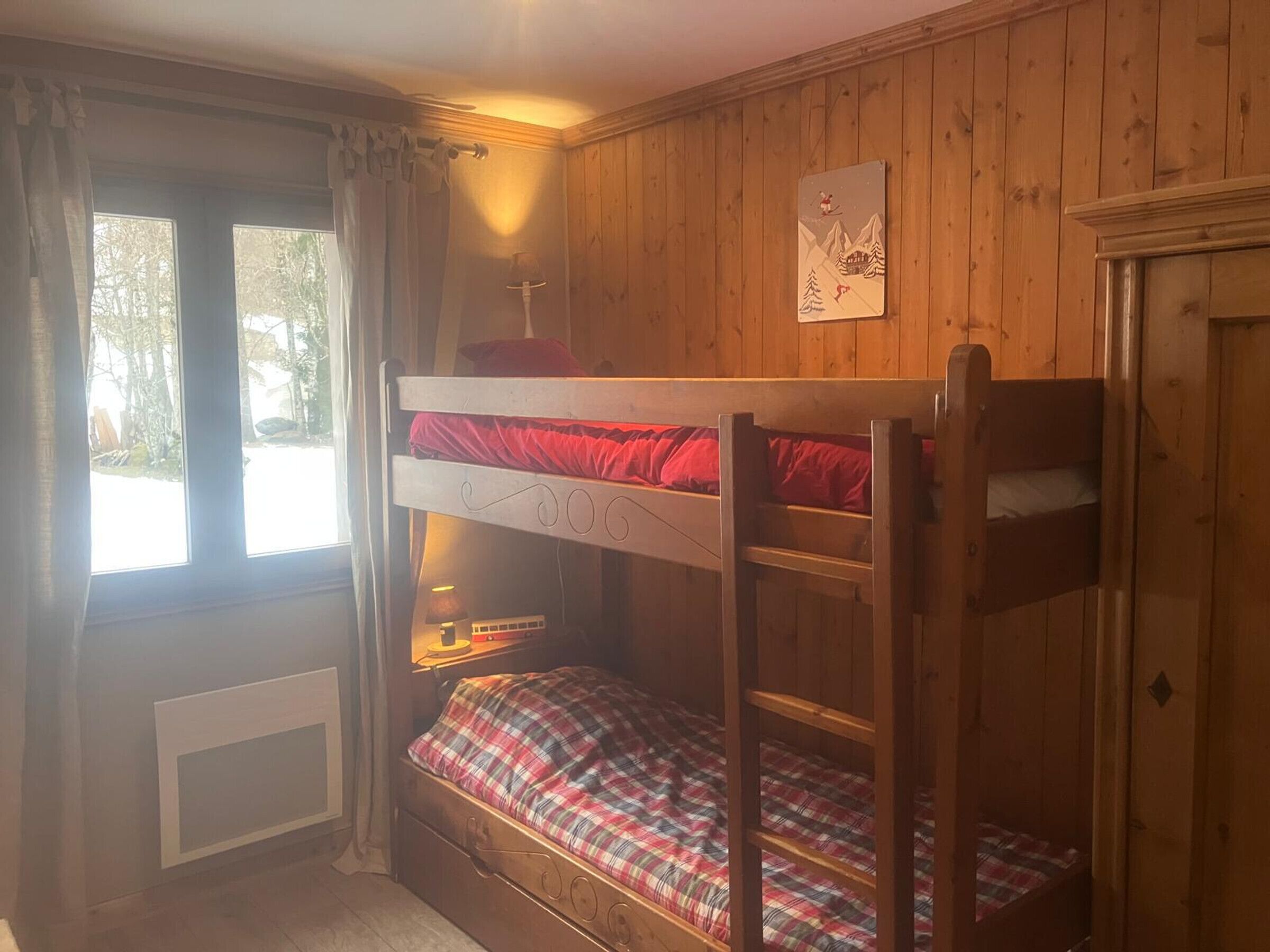 Habitació amb lliteres, parets de fusta, una finestra amb cortines i una taula petita amb una làmpada.