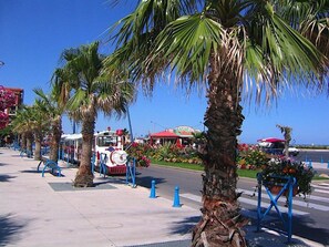 Eine Strandpromenade mit Palmen, einem Straßenkünstler und einem bunten Restaurant im Hintergrund.