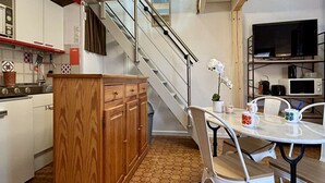 Une cuisine compacte avec des meubles blancs, un meuble en bois, une table à manger avec des chaises, un four à micro-ondes et un téléviseur posé sur une étagère.