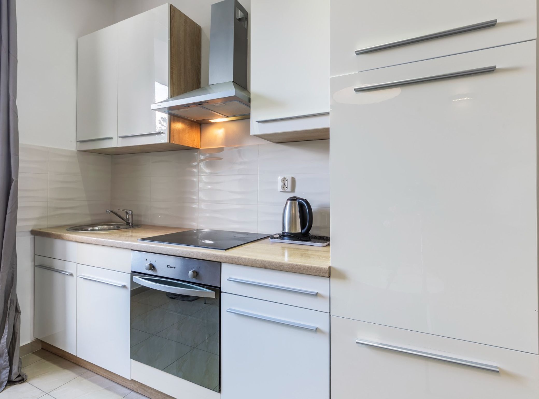 A modern kitchen with white cabinets, a stainless steel sink, a built-in oven, and a kettle on the countertop.
