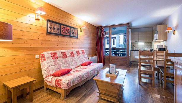 Um quarto aconchegante com piso de madeira, um sofá com capa estampada, uma pequena mesa de madeira e vista para uma varanda e área de cozinha.