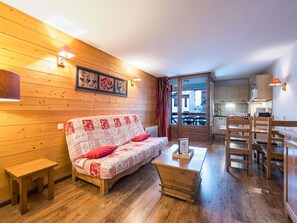 A cozy room with a wooden floor, a sofa with a patterned cover, a small wooden table, and a view of a balcony and kitchen area. - Cozy studio in Brides-les-Bains, near thermal baths and gondola, includes parking (Brides-les-Bains)