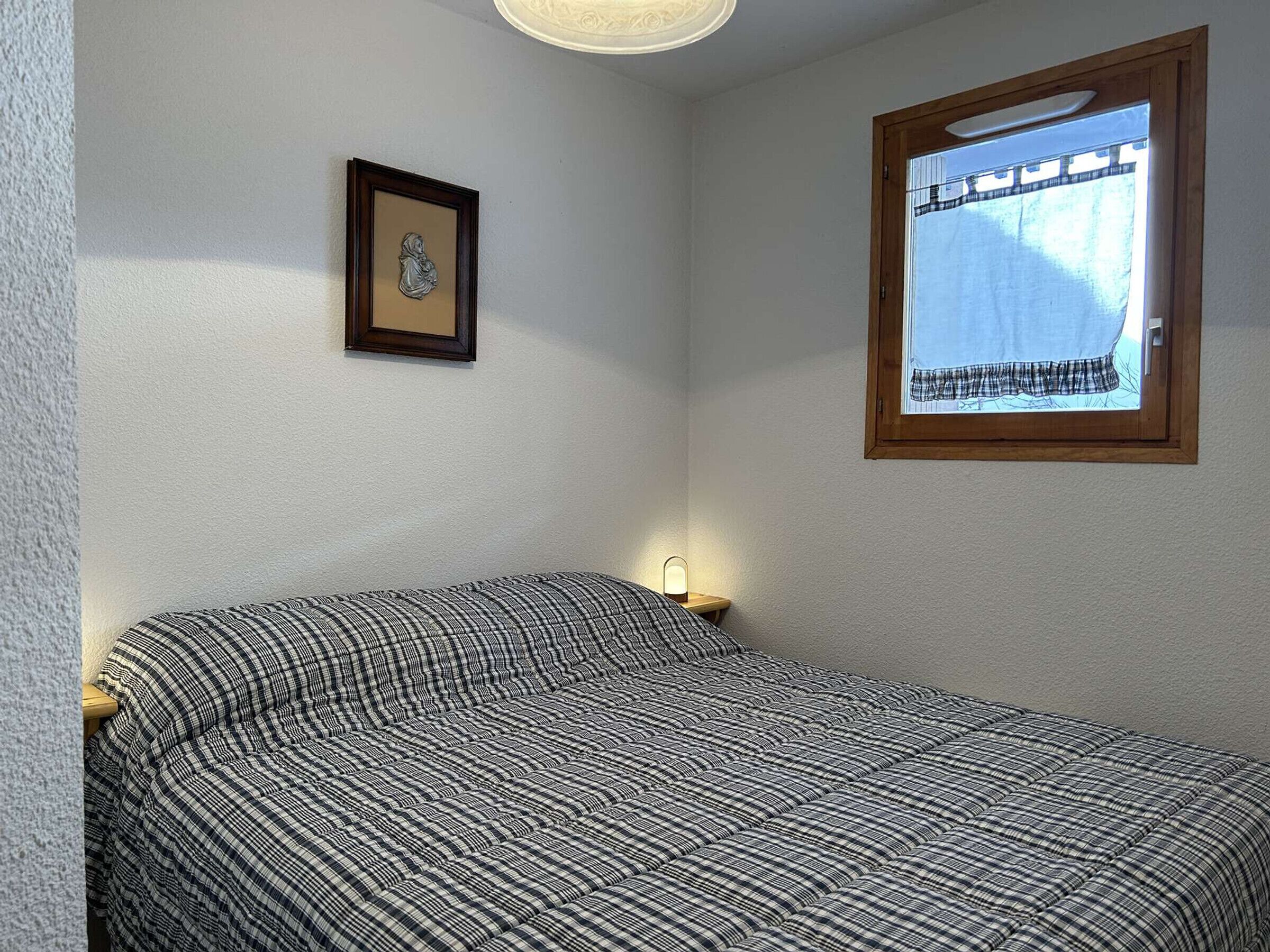A bedroom with a bed, a framed picture on the wall, a bedside lamp, and a window with a view of trees.