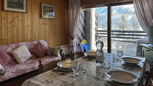 A cozy room with wooden walls, a sofa with floral cushions, a dining table set with plates and glasses, and a window with a view of snow-covered trees. - Cozy Apartment for 4 with Balcony and Parking in Montgenèvre (Montgenèvre)