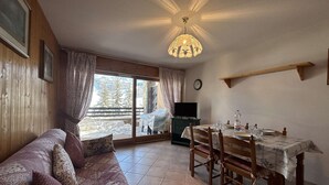 A cozy living room with a sofa, dining table, and chairs. A television is on a stand, and a window offers a view of a snowy landscape. - Cozy Apartment for 4 with Balcony and Parking in Montgenèvre (Montgenèvre)