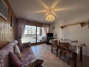 Un saló acollidor amb un sofà, taula de dinar i cadires. Una televisió reposa sobre un suport, i una finestra ofereix vista a un paisatge nevós.