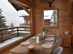 A wooden dining table set for four with a view of snow-covered trees and a cabin in the background. - Cozy Chalet in Montgenèvre with WIFI and Fireplace (Montgenèvre)