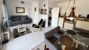 A modern living room with a grey sofa, a black armchair, a coffee table, and a dining area with a glass table and chairs. There is a kitchen area with a wooden cabinet and a red hanging light. - Spacious apartment 5 min from beach, 2 bedrooms, terrace, parking, WIFI (Roses)