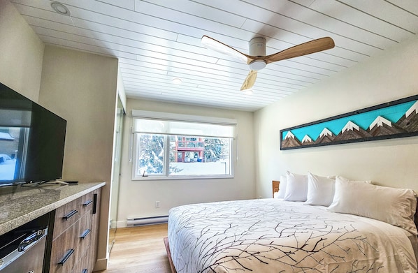 A bedroom with a bed, a ceiling fan, a window with a view, and a mounted artwork.