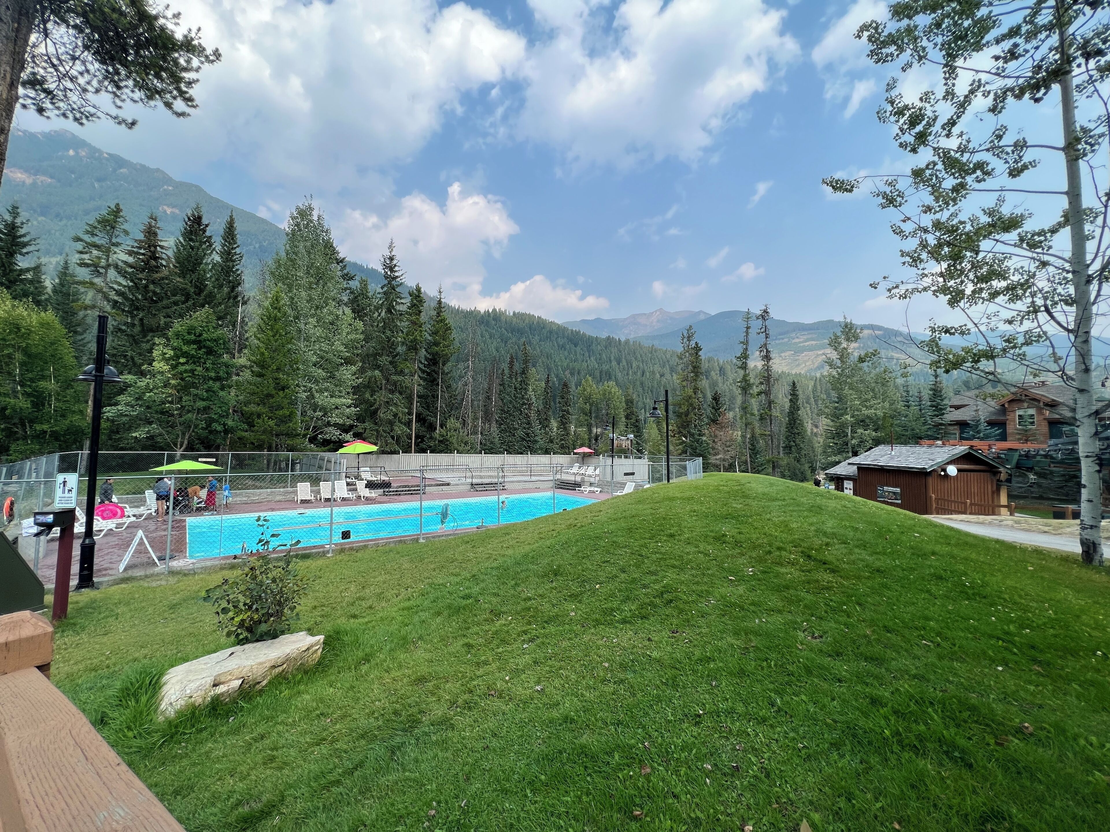 A swimming pool surrounded by a fenced area with lounge chairs and umbrellas, set against a backdrop of mountains and trees.