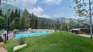 A swimming pool surrounded by a fenced area with lounge chairs and umbrellas, set against a backdrop of mountains and trees.