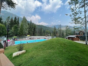 A swimming pool surrounded by a fenced area with lounge chairs and umbrellas, set against a backdrop of mountains and trees.