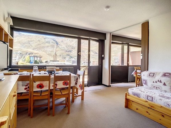 A compact room with a dining table and chairs, a sofa, and large windows offering a mountain view. - Bright Apartment · Ski-in/Ski-out · Terrace & Ski Locker (Les Menuires)