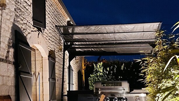 Een terras buiten met een stenen muur, een pergola, een barbecue en een eettafel met stoelen.