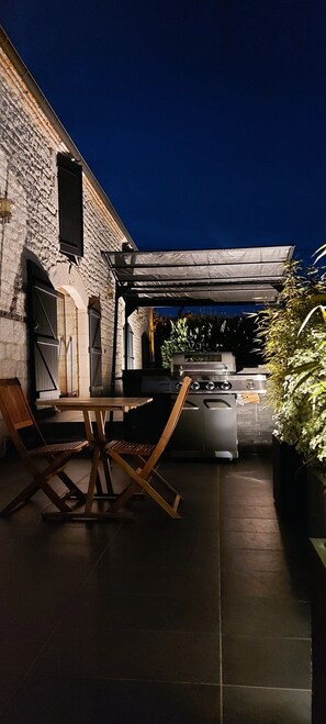 Une terrasse extérieure avec un mur en pierre, une pergola, un barbecue et une table à manger avec des chaises.
