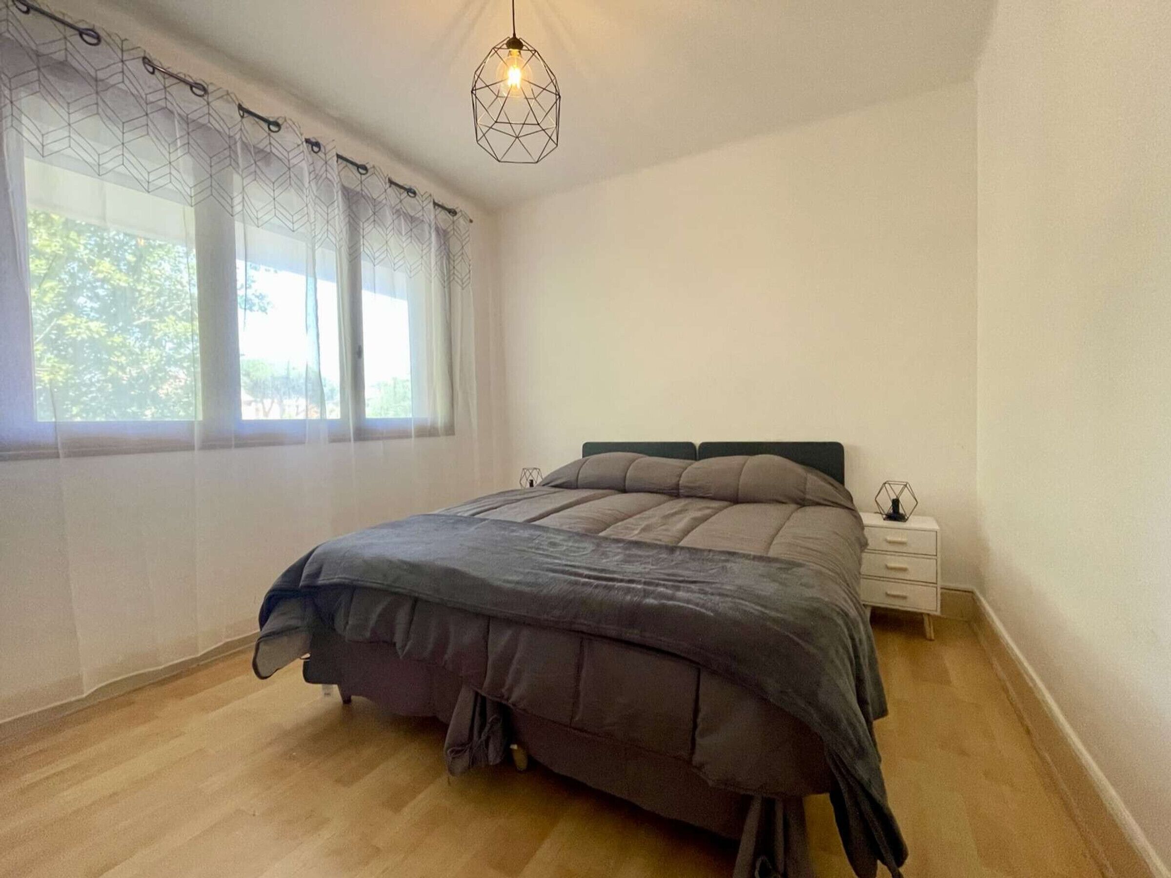 Un dormitorio con cama, mesita de noche, lámpara colgante y ventana con cortinas traslúcidas.