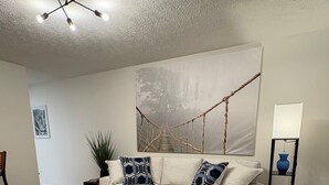 Una sala de estar con un sofá color beige, cojines estampados, una mesa de centro de madera, un cuadro abstracto grande, una lámpara de pie y una planta en maceta.