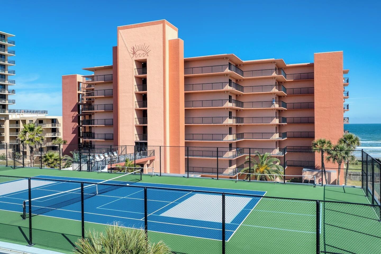 A multi-story hotel with balconies, a tennis court, and a view of the ocean.