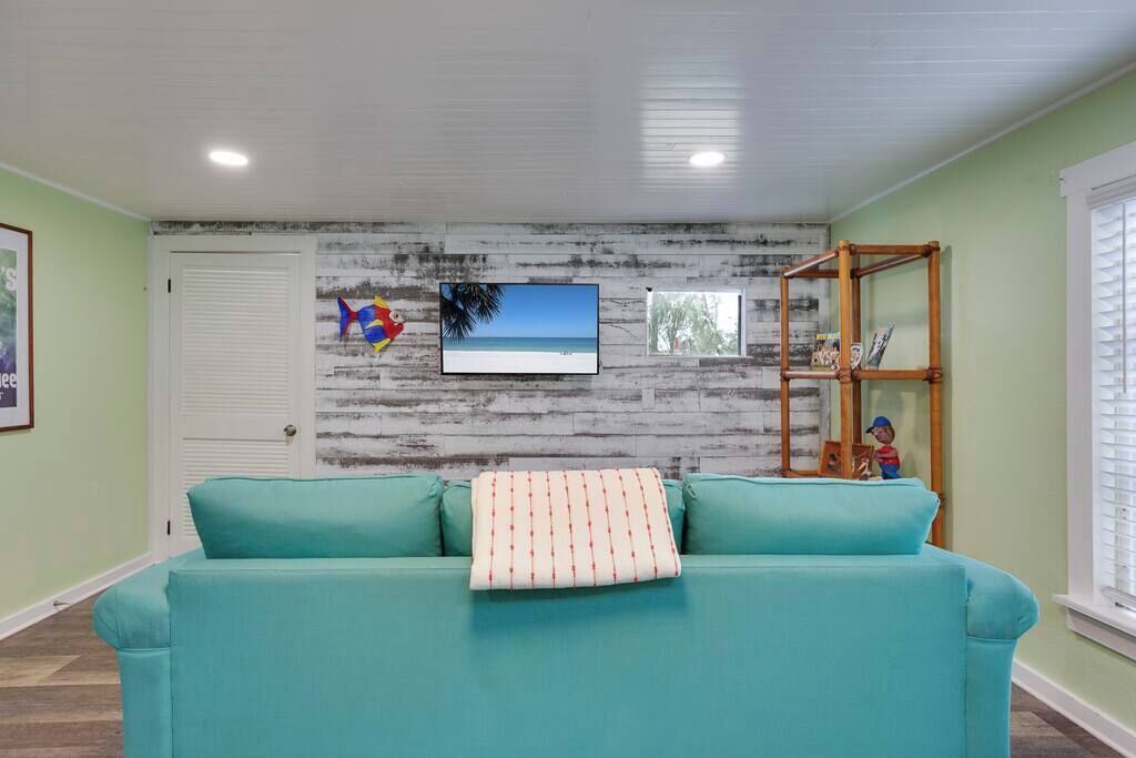 A teal sofa with a patterned pillow, a television mounted on a distressed wall, and a wooden shelf with decorative items.