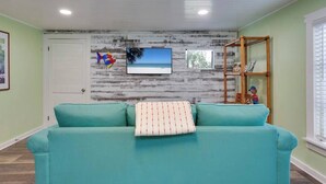 Um sofá azul-esverdeado com almofada estampada, uma televisão presa a uma parede desgastada e uma prateleira de madeira com objetos decorativos.
