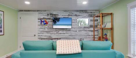 A teal sofa with a patterned pillow, a television mounted on a distressed wall, and a wooden shelf with decorative items.