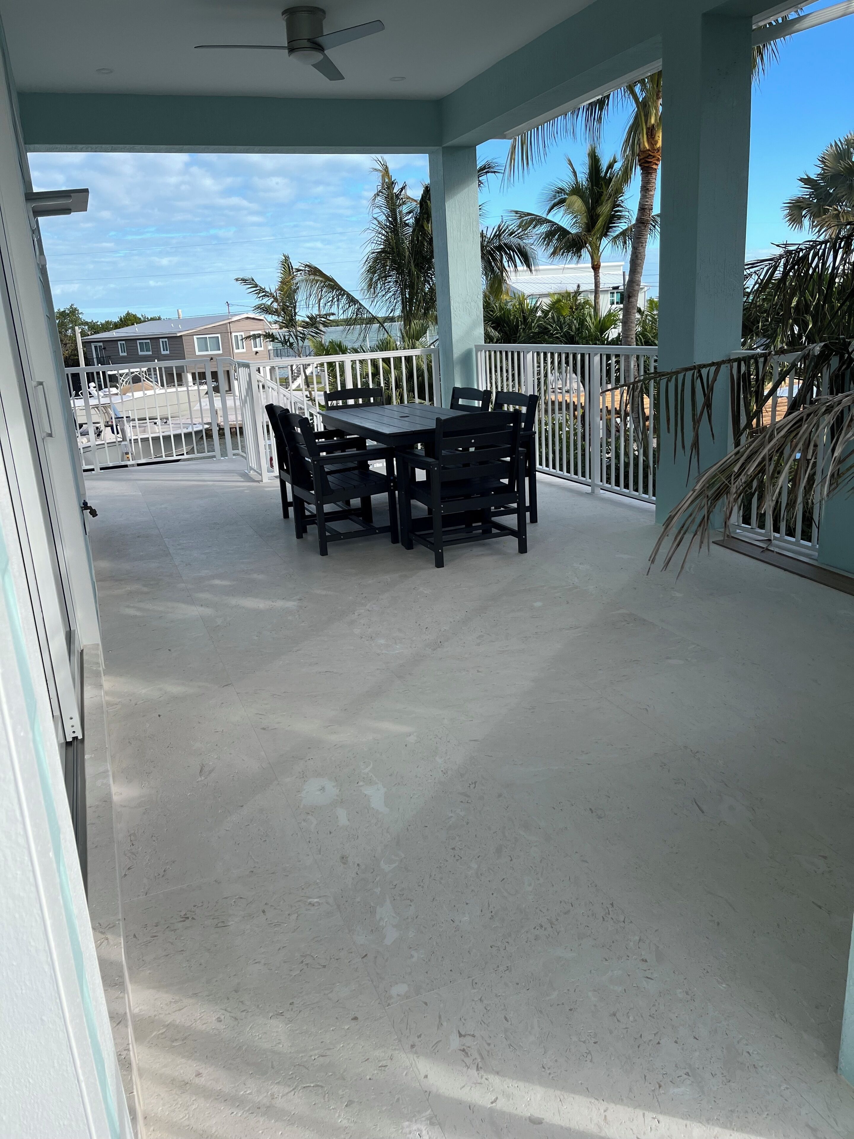 Eine überdachte Terrasse mit Tisch und Stühlen, umgeben von einem weißen Geländer, Palmen und Blick auf das Meer.