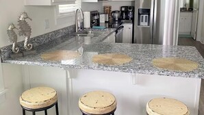 Una cucina con mobili bianchi, un piano di lavoro in granito, un frigorifero in acciaio inossidabile e tre sgabelli in legno.