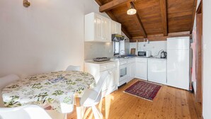 Une cuisine compacte avec des meubles blancs, une cuisinière, un four à micro-ondes et un réfrigérateur. Le plafond est en bois et il y a un petit coin repas avec une nappe à motifs floraux.