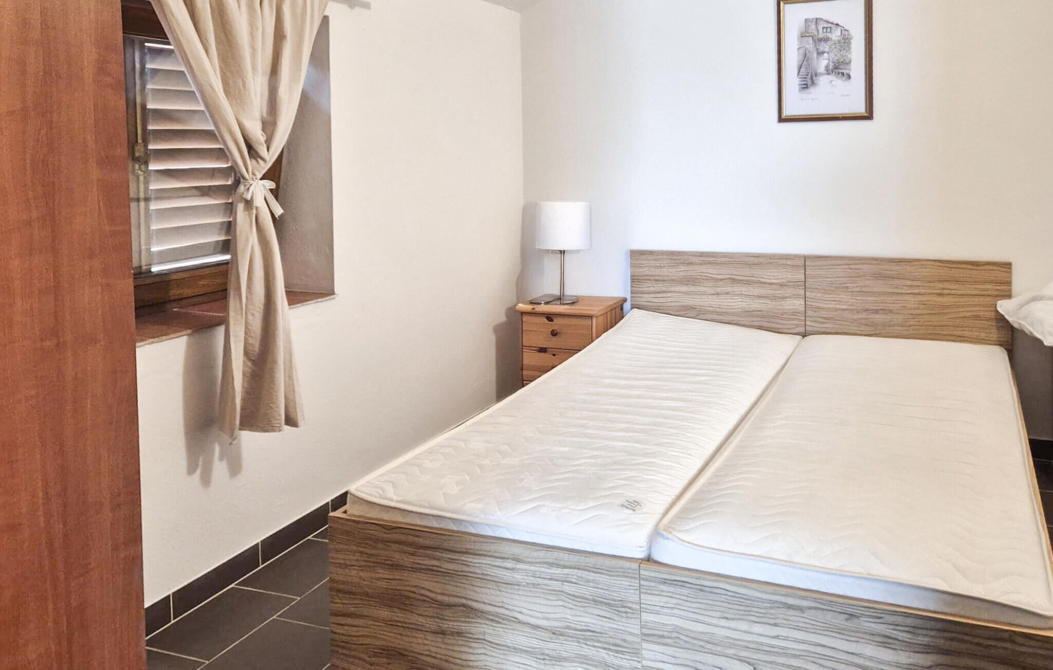Un dormitorio con cama de madera, una mesita de noche con lámpara, una ventana con cortinas y un cuadro enmarcado en la pared.