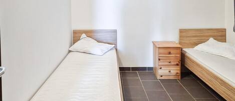 A small, clean room with two beds, a wooden nightstand, and a tiled floor.