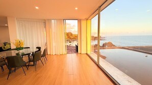 Un cuarto amplio con piso de madera, una mesa de comedor con sillas y un ventanal con vistas al mar y a la costa rocosa.
