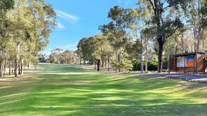 Camp de golf amb una construcció de fusta a la dreta, envoltat d'arbres i un cel serè.