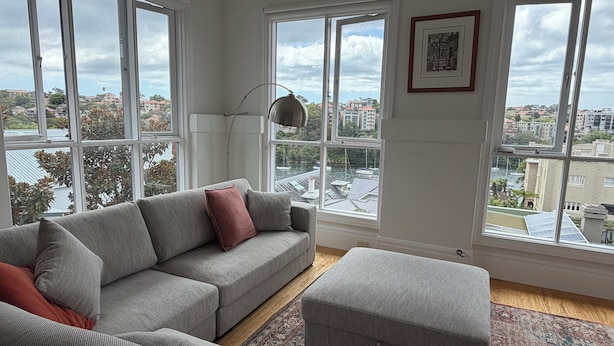 Uma sala de estar moderna com um sofá cinza, um banquinho e um abajur. Janelas amplas oferecem vista para um horizonte urbano e um corpo d'água.