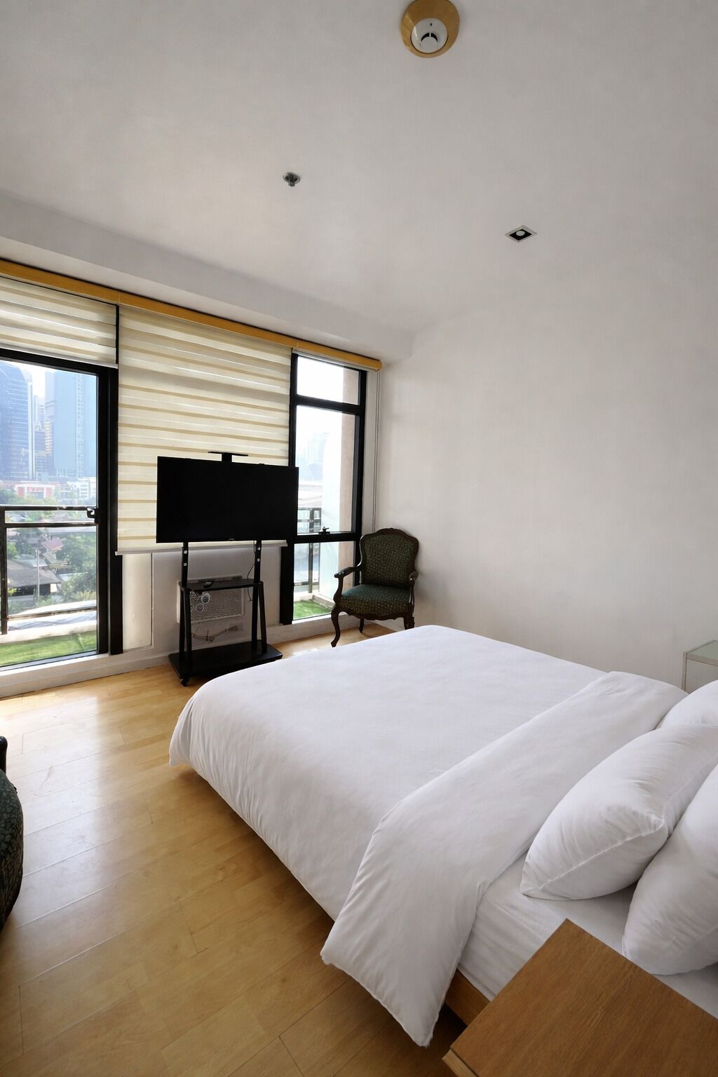 Un dormitorio con una cama, una silla, un televisor sobre un mueble bajo y una ventana con persianas.