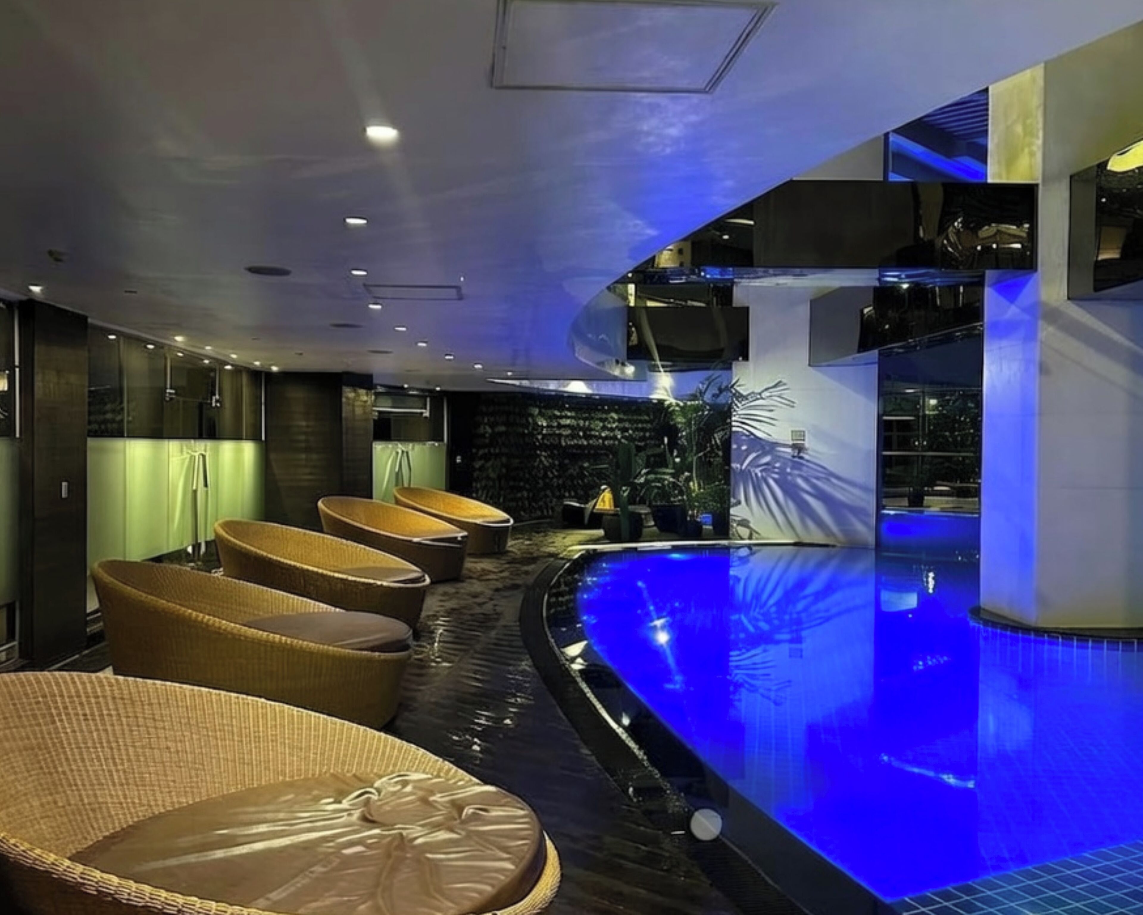 A modern indoor pool area with a curved design, featuring wicker lounge chairs and a blue-lit pool.