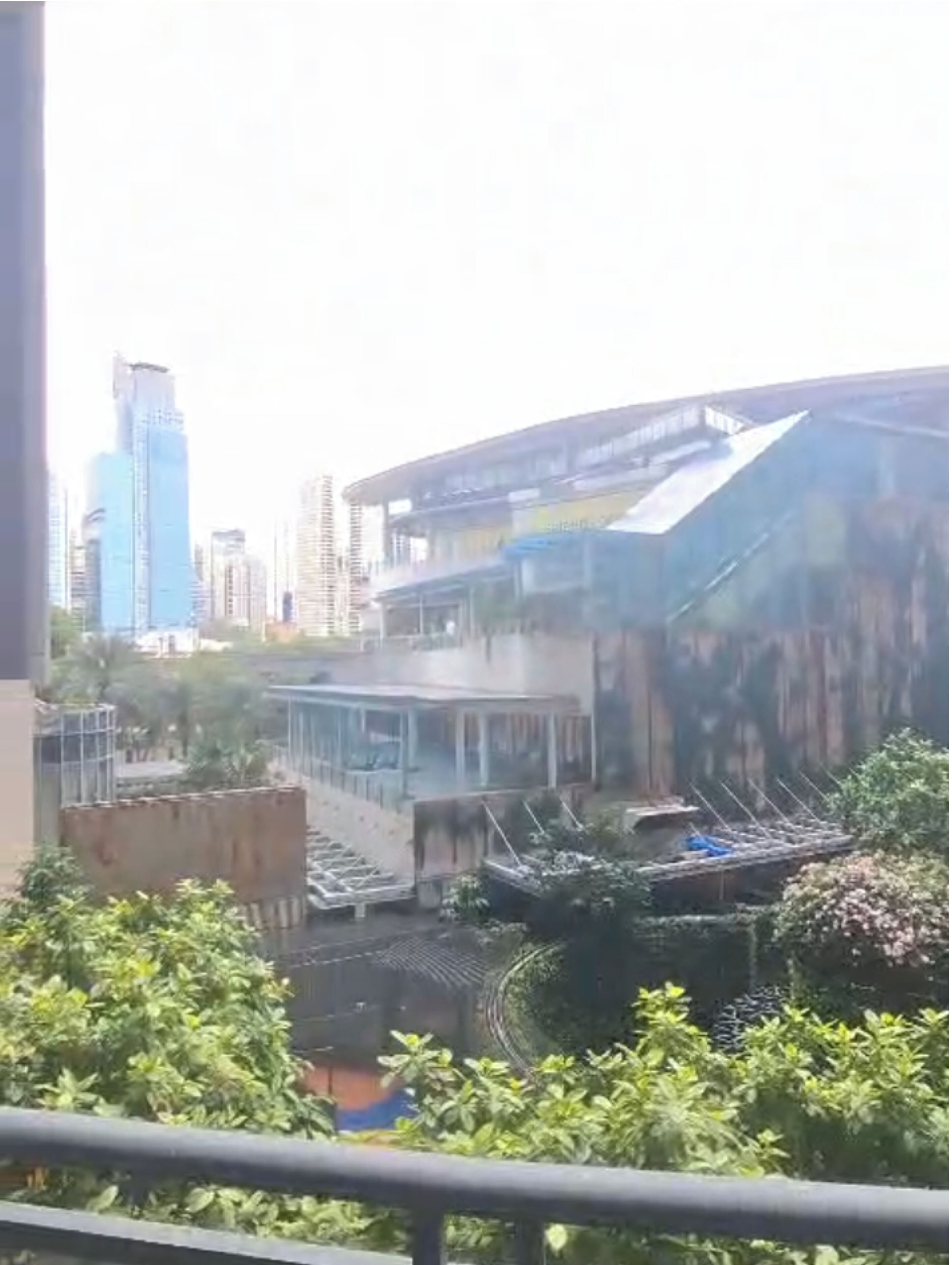 A view of a modern building with a curved facade and a flat roof, surrounded by greenery and other buildings in the background.