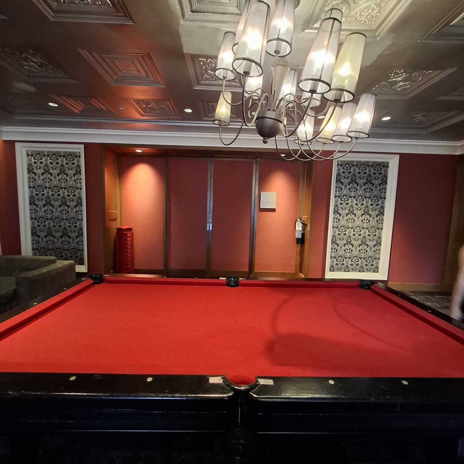 A billiards table with a red felt surface, surrounded by patterned wallpaper and a chandelier.