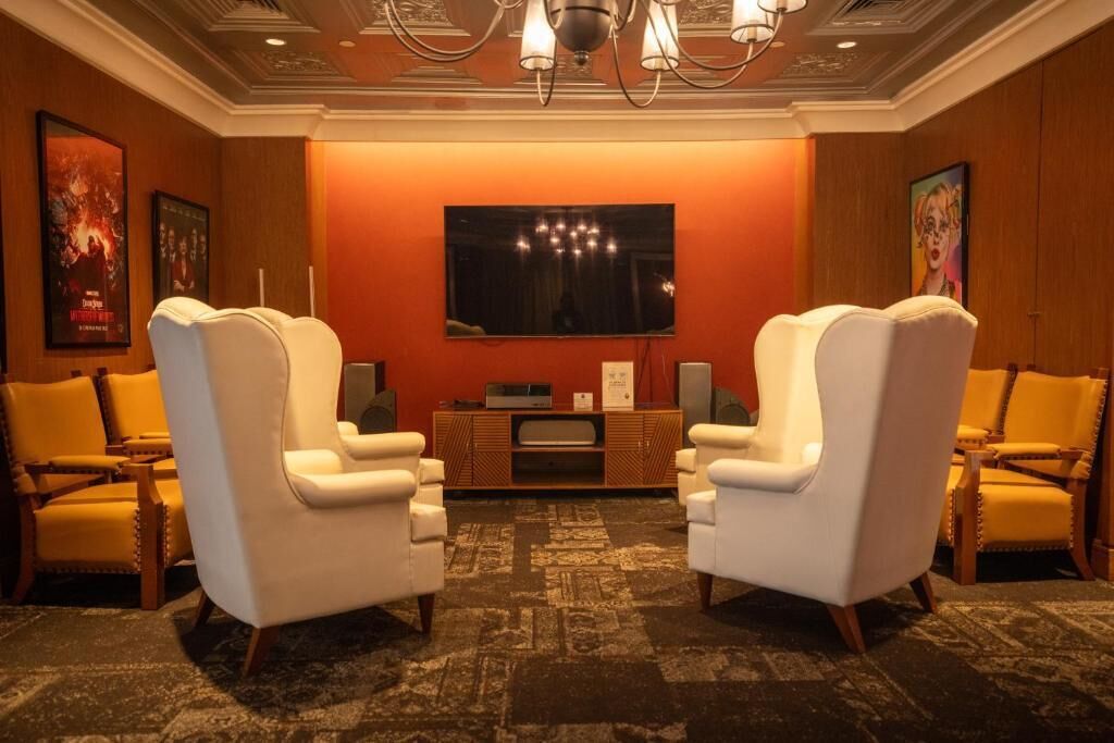 A hotel room with a large flat-screen TV mounted on the wall, two white armchairs, a wooden side table with a flat-screen TV, and a patterned carpet.
