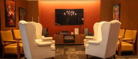 A hotel room with a large flat-screen TV mounted on the wall, two white armchairs, a wooden side table with a flat-screen TV, and a patterned carpet.