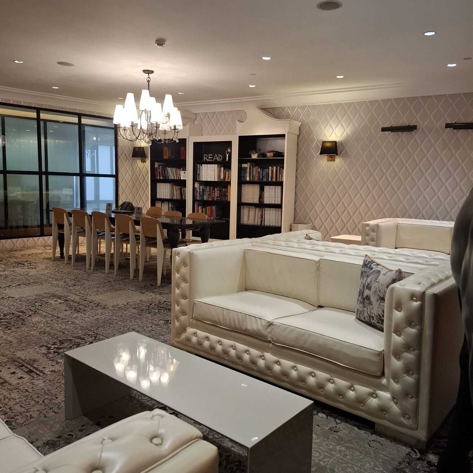 A modern living room with a white leather sofa, a glass coffee table, and a chandelier. There are bookshelves filled with books and a dining area with chairs and a table.