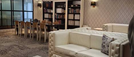 A modern living room with a white leather sofa, a glass coffee table, and a chandelier. There are bookshelves filled with books and a dining area with chairs and a table.