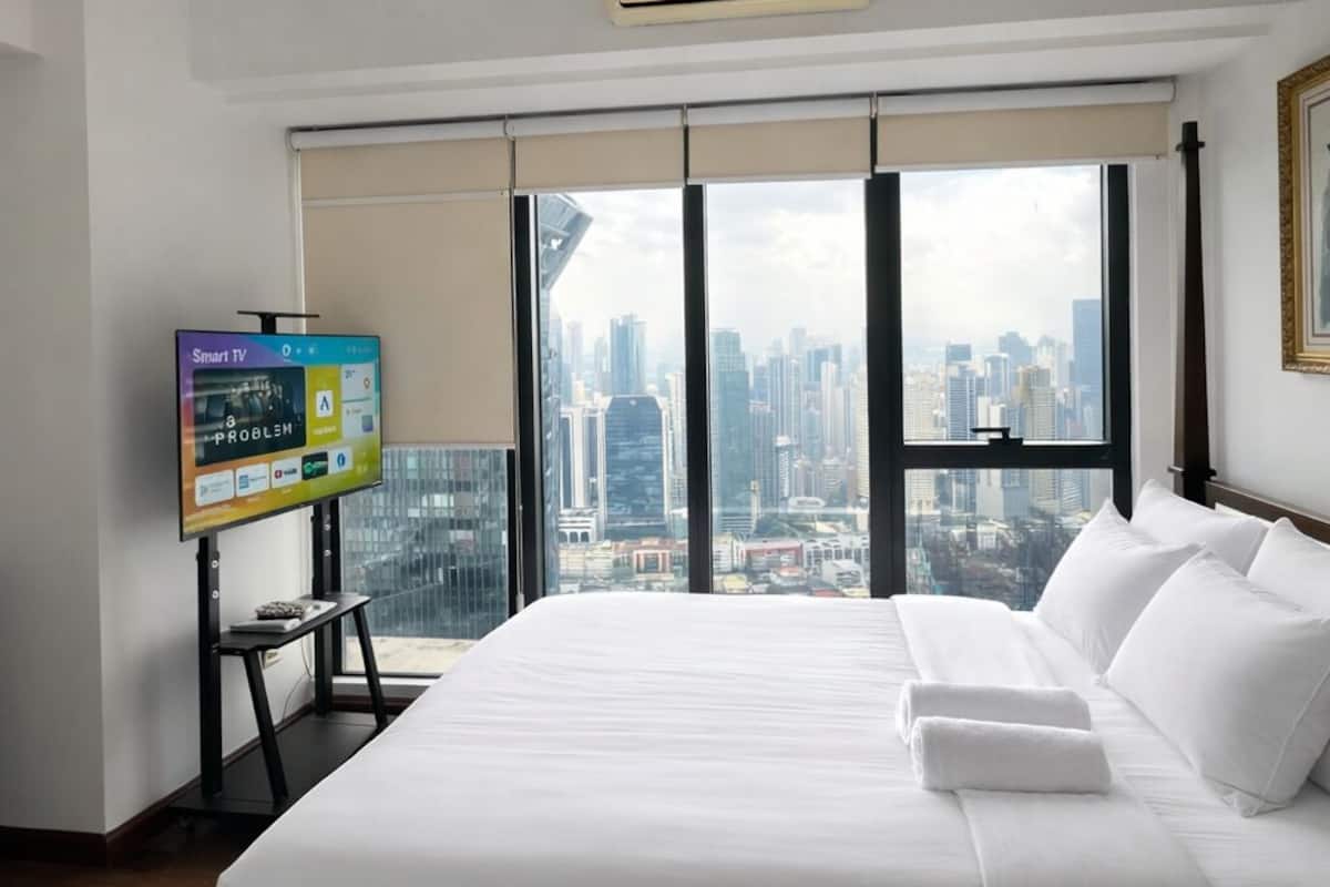 A hotel room with a large bed, a TV on a stand, and a city view through floor-to-ceiling windows.