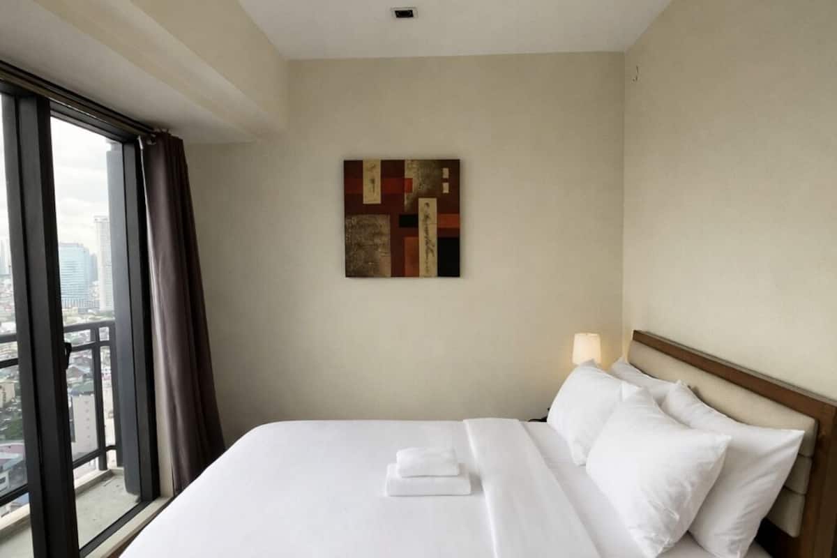 A hotel room with a large bed, a window with curtains, a ceiling light, and a wall-mounted artwork.